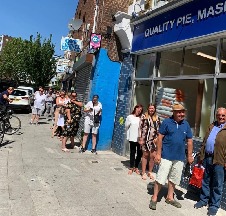 People wait in pairs in line outside a shop with a blue storefront on a sunny day.