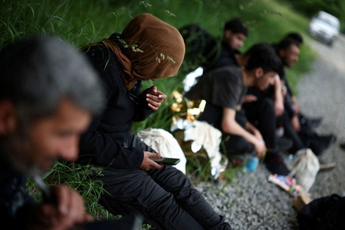 Migrants are seated on the ground in a forest near Grudki, Poland, after crossing the Belarusian-Polish border. They appear to be resting, with one person holding a phone. 