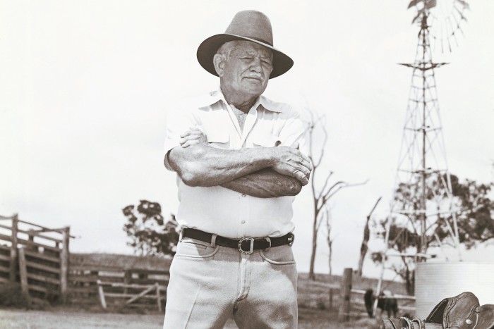 A black and white image showing RM Williams standing with his arms folded next to a saddle