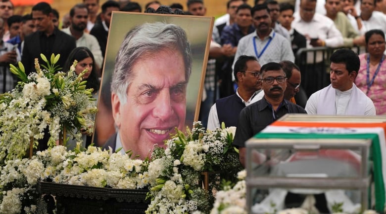 A crowd of people stand near a giant portrait showing a close-up of a smiling older man's face. The picture is surrounded by white flowers. On the right side of the image, a clear casket draped in a flag is visible. 