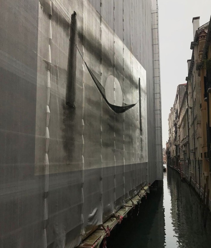 A photograph shows a narrow stretch of canal in Venice with buildings rising to one side and on the other side buildings clad in scaffolding and material covering