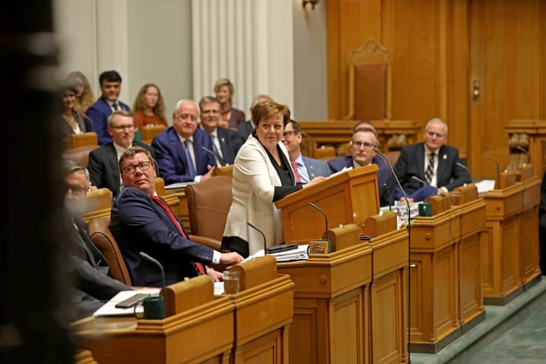 A woman in a white blazer stands at a podium among other people in dress clothes beside and behind her in a formal legislature setting