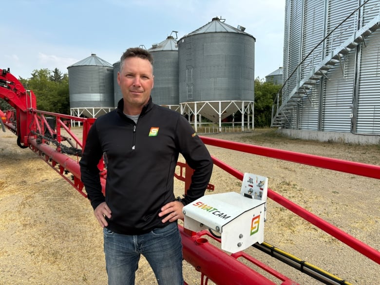 Derek Rude stands next to white box with camera attached to farm machinery, with metal silos in background