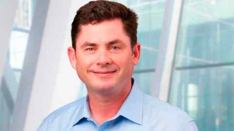 Brendan Horgan, wearing a light blue shirt, smiles while standing in front of a glass background.