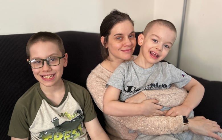 A boy with glasses smiles while sitting on a dark couch next to his mother, who holds his younger brother on her lap.