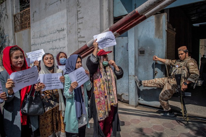 Women in Afghan clothing walk together holding up sheets of printed paper; a soldier looks on, leaning on his rifle