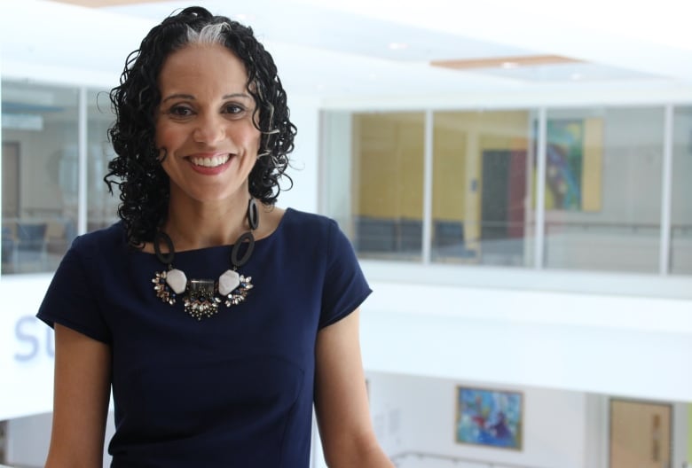 A woman with black curly hair and a white stripe, smiling, wearing a dark blue top and white beaded necklace. 