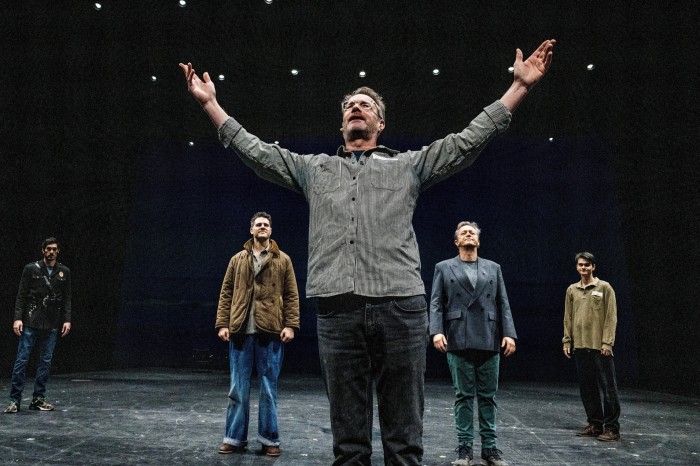 A man stands on stage, his arms  held up to the sky. Four other men stand behind him