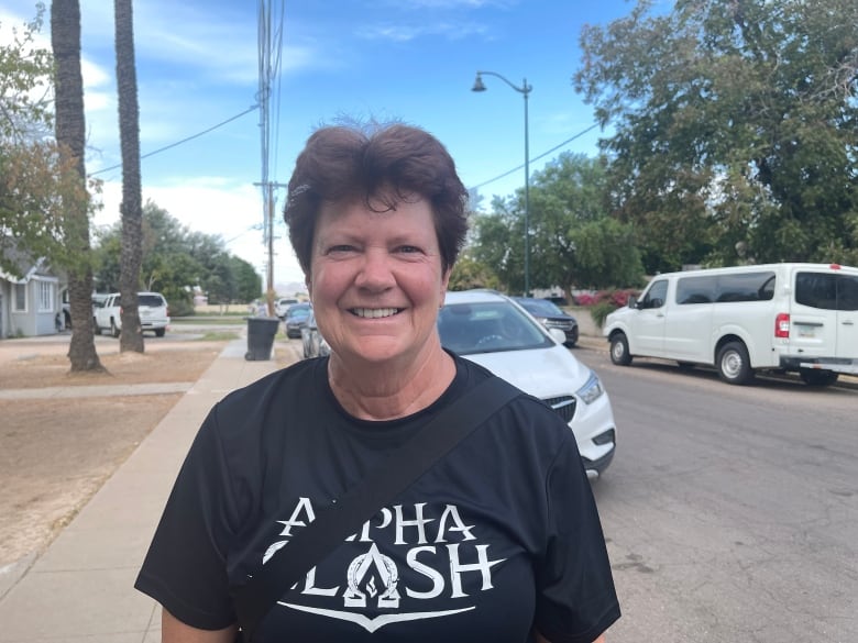 A woman with cropped hair, standing on a neighbourhood street, smiles.