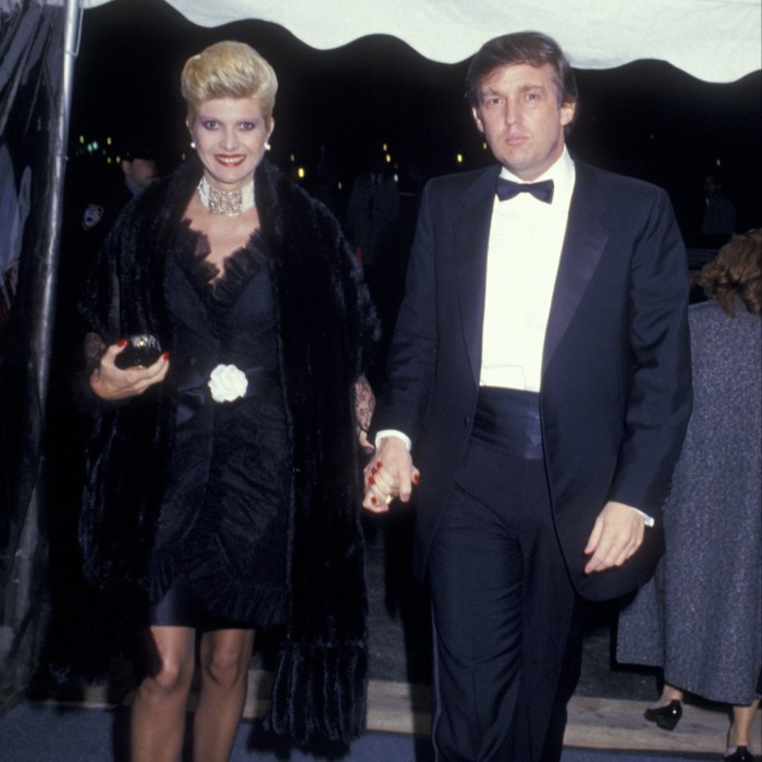 A smiling woman with blonde hair, wearing a black evening gown and stole, holds the hand of a man in a tuxedo as they arrive at an event