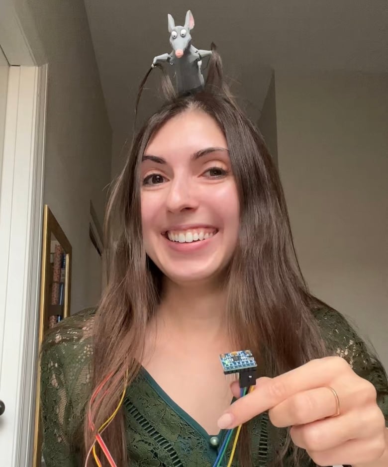 A woman holds up wiring while a robotic rat sits on her head. 