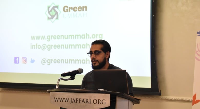 A man with a beard and glasses speaks at a podium. A slide in the background says Green Ummah, www.greenummah.org.