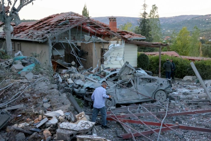 A person examines the debris at the site of an Israeli air strike. The image shows a heavily damaged house and a wrecked car amid the rubble.