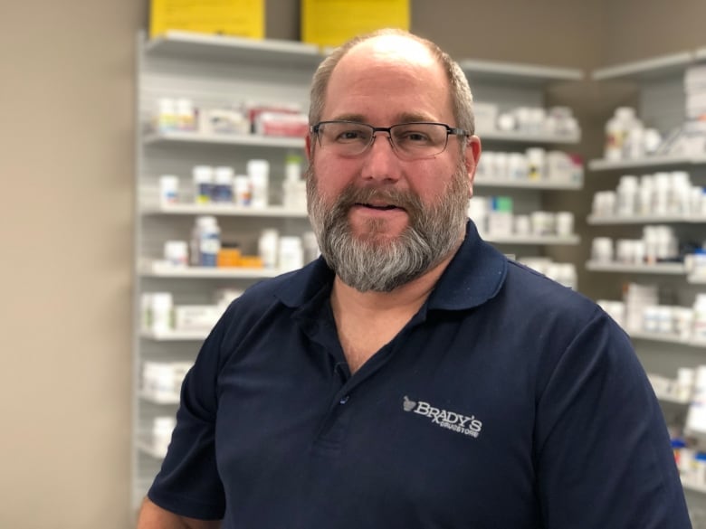 A man in a blue polo that reads "Brady's Drugs" stands in front of a shelf of medications