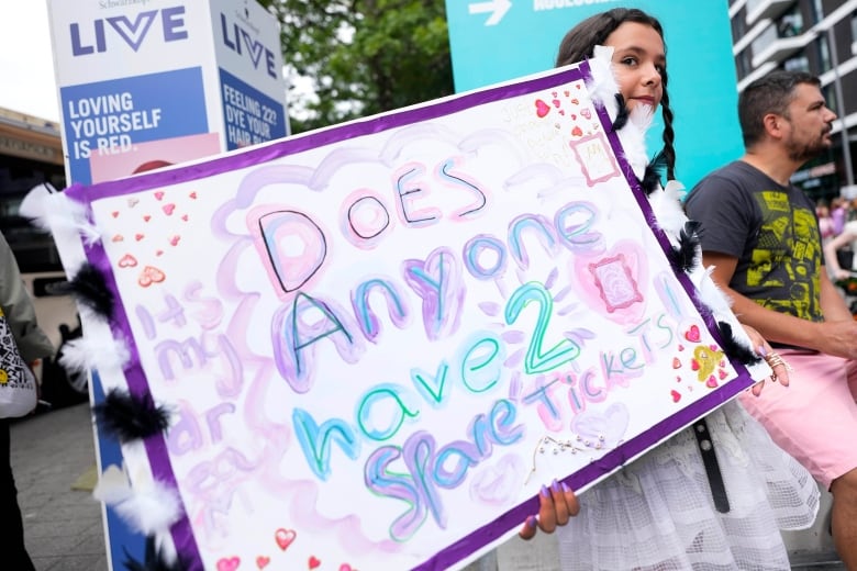 A girl holds a hand-made sign that says "Does anyone have 2 spare tickets?"