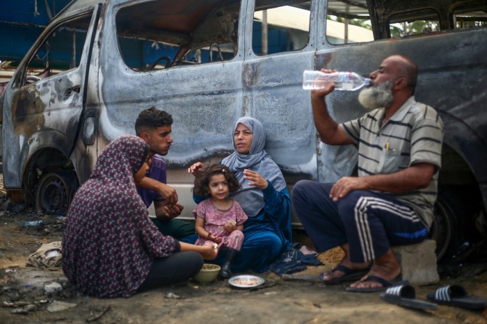 A group of people share a meal beside a burnt out vehicle