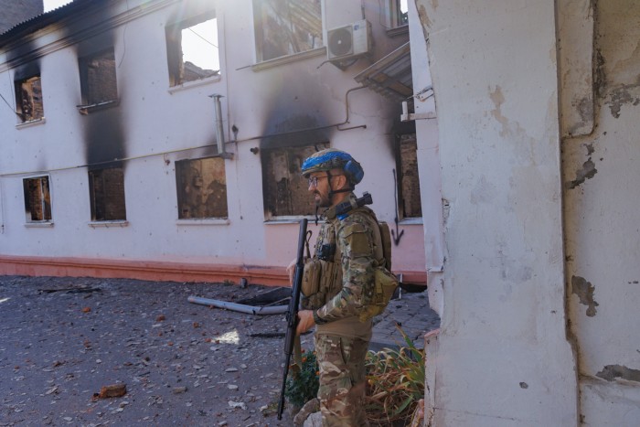 Ukrainian military man with a Kalashnikov rifle stands near destroyed building 