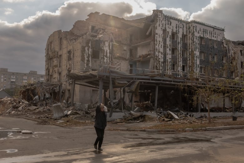 A lone person is shown on an empty road in front of a multistorey low rise building that has been destroyed and hollowed out, with debris on the ground nearby.