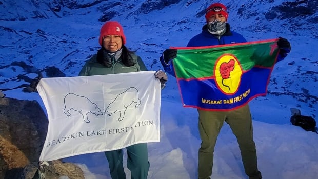 Two people raise First Nations flags on a snowy mountaintop.