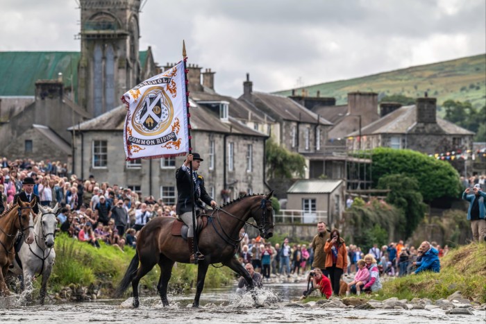 A man is riding a horse through shallow river, holding a large white flag. A large crowd has gathered on both sides of the river to watch. In the background, there are stone buildings