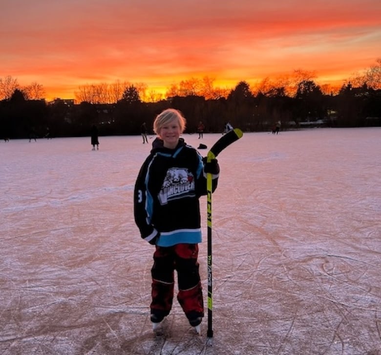 A smiling boy wearing a hockey jersey and holding a hockey stick stands in his skates on a frozen outdoor rink as the sun sets. 