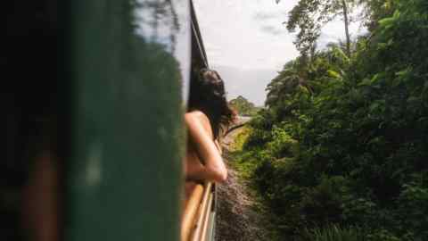 A woman in white trousers and gauzy white shirt shades her eyes as she looks out from a train carriage