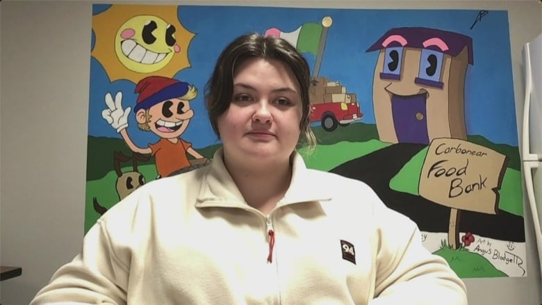 Woman with brown hair pulled back, wearing a white sweater. She's seated and a mural behind her shows a sign that says "Carbonear Food Bank" as well as a cartoon sun with eyes that's smiling.