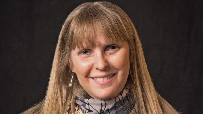 A smiling woman with long blonde hair stands against a black background.