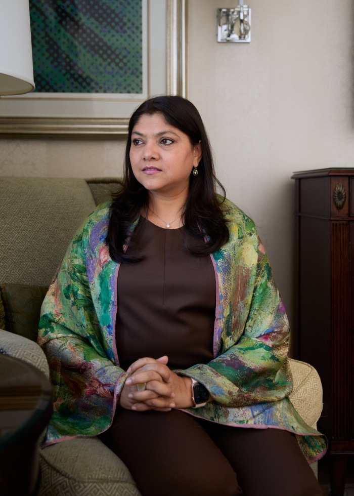 A woman sits in an armchair, wearing a colorful shawl over a dark brown outfit. Her hands are clasped in her lap, and she gazes thoughtfully to the side