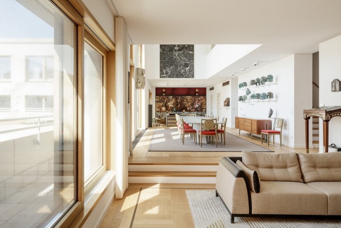 A living and dining area in a luxury home or apartment. On the left are large floor-to-ceiling windows. In the foreground, there is a beige sofa with soft cushions. At the back is a raised dining area accessible by a few steps