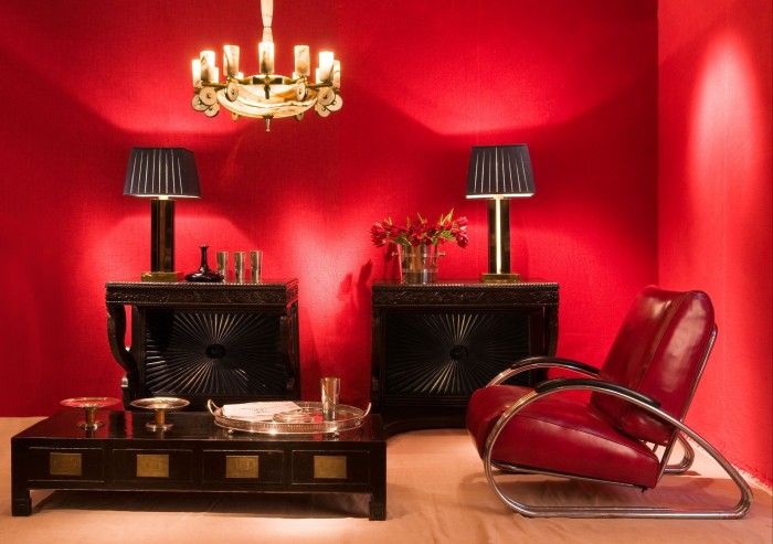 A room with bright-red walls displays a number of furniture pieces: a red-leather armchair with a tubular chrome frame; a Modernist gold-toned chandelier; a low, dark-wood coffee table with silver trays and bowls on top; and a pair of engraved, dark-wood sideboards 