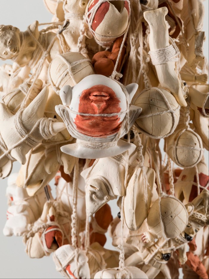 A series of white ceramic and porcelain shapes, including one inscribed with a human face, are bound into a hanging sculpture by thick white string.