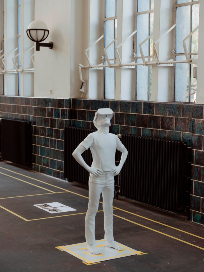 White plaster sculpture on the floor of an art studio, depicting a boy with his hands on his hips, staring upwards, wearing VR goggles