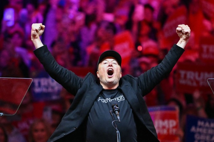 Elon Musk raises both arms in the air as he steps onstage at a Trump rally