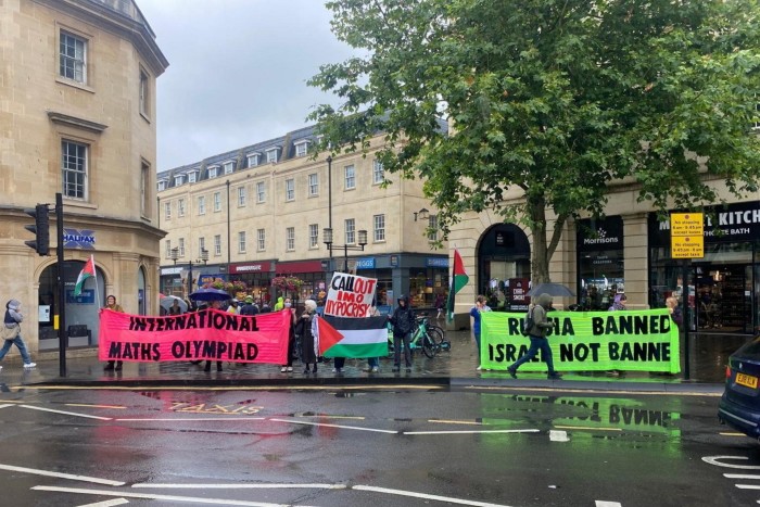 Protesters are standing in the street holding banners during the IMO final. One pink banner reads “International Maths Olympiad,” while another says “Call Out IMO Hypocrisy” with a Palestinian flag design. A green banner reads “Russia Banned, Israel Not.” 