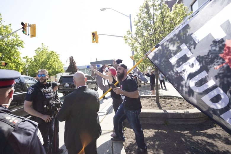 Police stand in front of a bearded man waving a large flag - the text, 'F--k Trudeau,' is only half visible within the image. 