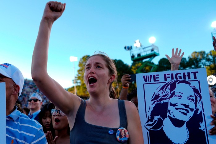 Suffused in blue light, a woman in a sleeveless blue top raises her fist to the sky. She wears a Kamala Harris badge. Next to her is a poster, in blue, of Harris. On the other side stands a man in a blue polo shirt and white Kamala Harris baseball cap