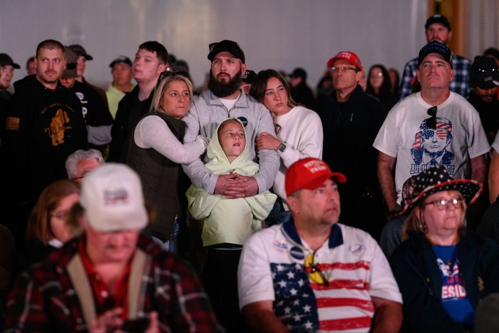 An audience of mainly white people, many of them wearing red baseball caps, heads turned attentively towards the stage. In the centre of the picture, a woman and two girls hug a man in what looks like a gesture of hope or fear