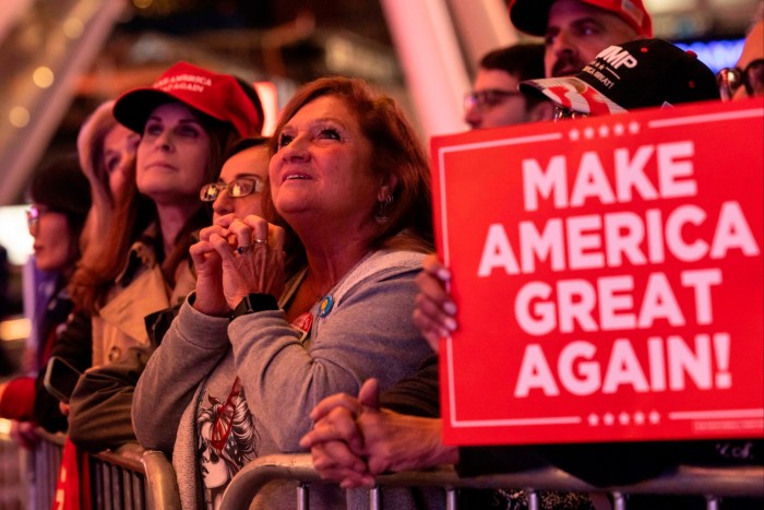 Suffused in red light, a group of women at the front with a few men behind stand rapt with attention at crowd control barriers. One of them holds a Make America Great Again placard, also in red 