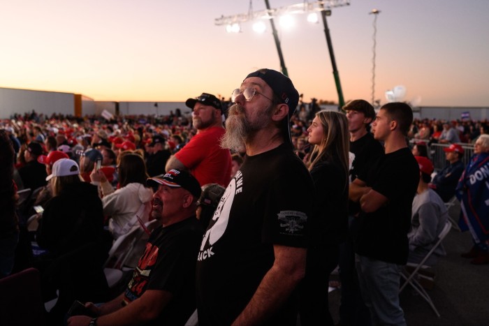 Standing outdoors at sunset, a group of people look expectantly in one direction. In the foreground are mainly white men in black T-shirts; further away are many others in red baseball caps