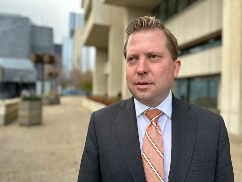 A man with blonde hair, wearing a black suit jacket and an orange tie, stands outside in front of a concrete building.