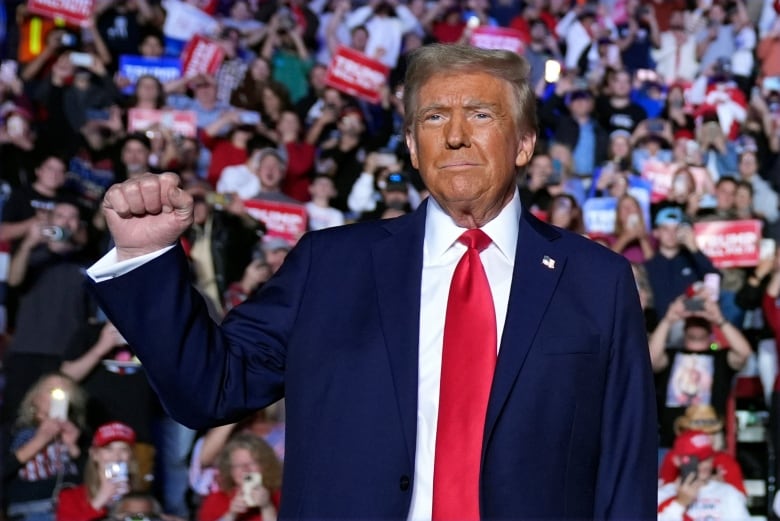 A man in a navy suit with a red tie raises his fist and smiles on stage at a political rally.