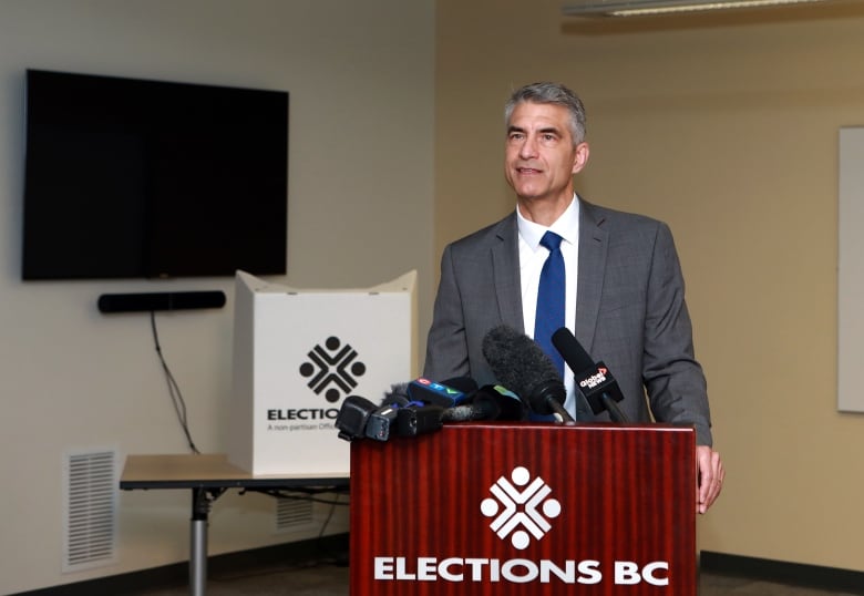 A man stands at a podium marked 'Elections BC' with a gaggle of microphones.