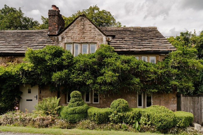 A stone cottage with a slate tiled roof, and trees and shrubs in the front garden