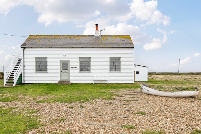 A small, white, one-story house with horizontal siding and a grey, shingled roof on a patch of grass, with a rocky and pebbly ground