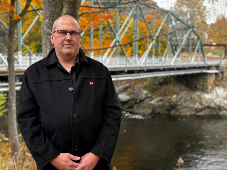 A man in a black jacket looks ahead at the camera, with a bridge and autumn leaves in the background.