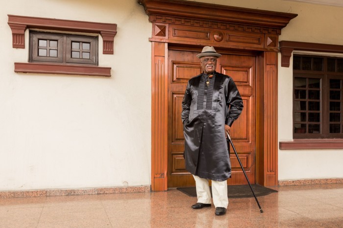 A man in a black robe and tan trousers holds a cane outside an elaborate wooden double door