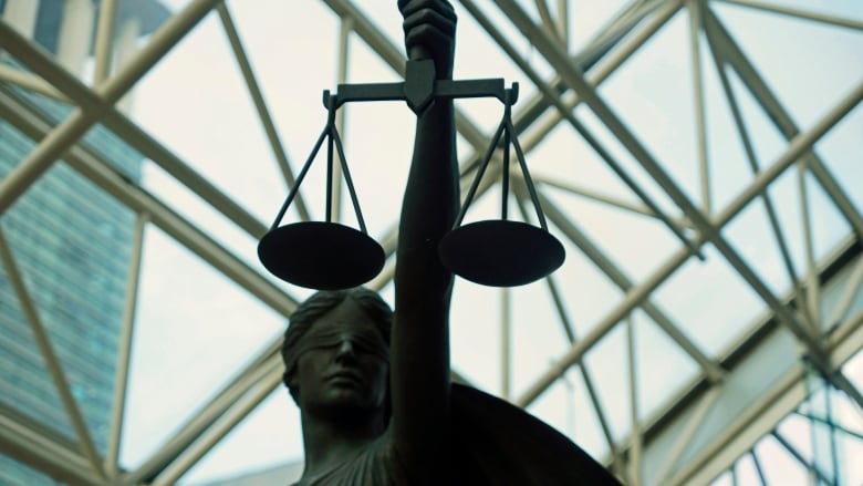 The statue of Themis, Goddess of Justice, is pictured beneath a glass roof. It is a blind woman holding up scales.