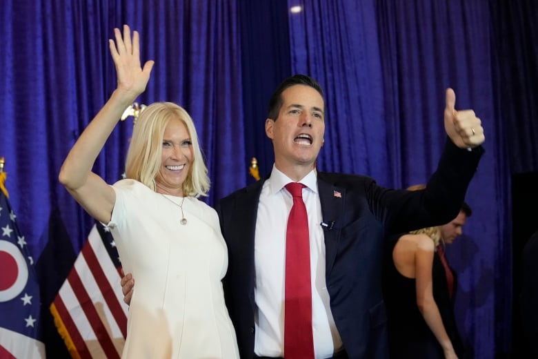 A cleanshaven dark haired man in a suit and a blonde woman wearing a white dress smile and raise their arms on a stage.