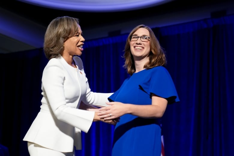 Two women, one with glasses and Caucasian and the other dark skinned, embrace on a stage while smiling.